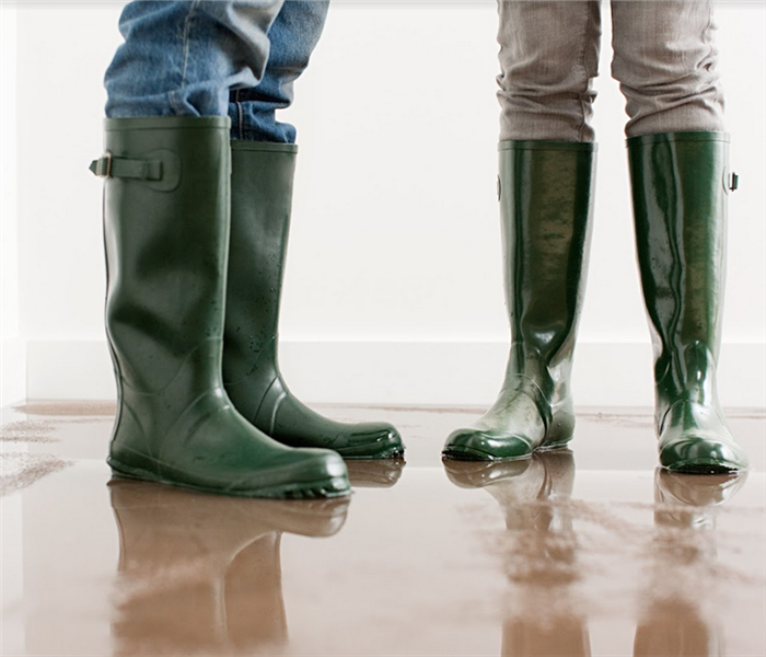 two people standing in rain boots in a flooded room with waterlogged carpet on the floor